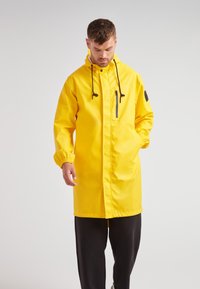 Žlutý voděodolný plášť do deště s kapucí, přední kapsou na zip, elastickými manžetami a bočními kapsami. Na rukávu je černé kulaté logo.