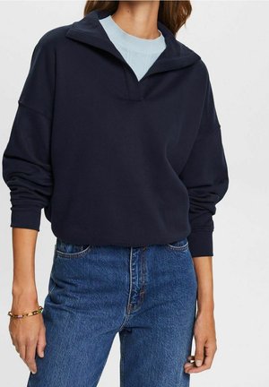Navyblauwe oversized pullover met een kwart ritssluiting en ribgebreide manchetten, gecombineerd met hoge blauwe jeans en een lichtblauw t-shirt eronder.