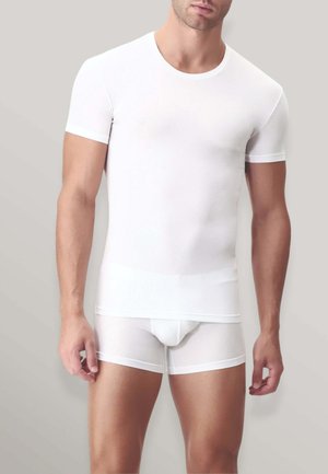 Modello maschile che indossa una maglietta bianca aderente a maniche corte e boxer bianchi su uno sfondo grigio chiaro uniforme.