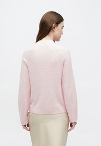 Svetloružový pletený sveter s guľatým výstrihom, raglánovými rukávmi a jemnou textúrou, v kombinácii s neutrálnou sukňou.