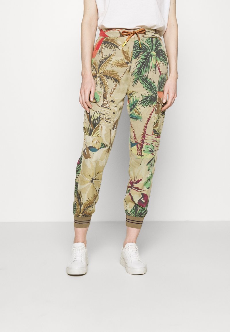 Personne portant un pantalon cargo beige à imprimé tropical avec des palmiers et des fleurs, assorti à des baskets blanches et un haut blanc.