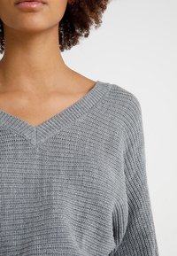 Femme aux cheveux bouclés portant un pull en maille gris clair à col en V, sur un fond blanc uni.
