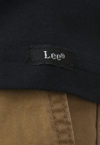 Țesătură neagră cu un etichetă ce arată "Lee" în text alb, cusută pe margine. Partea inferioară are un material de culoare bej, cu cusături vizibile.