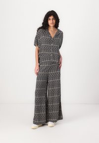 Svart og krem mønstret jumpsuit med korte ermer, dyp V-hals, vide bein og en avslappet passform, kombinert med lyse sko.