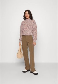 Pull en tons de rose avec des détails en franges, associé à un pantalon marron, des chaussures noires avec semelles blanches et un sac à main beige texturé.