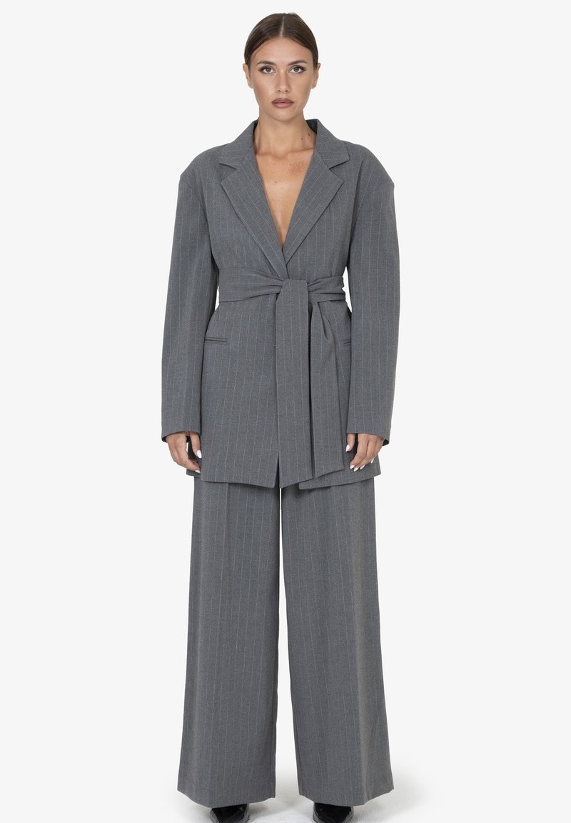 Vrouw die staat in een grijze gepindste strikblazer met riem, met bijpassende wijde broek en zwarte schoenen, tegen een witte achtergrond.