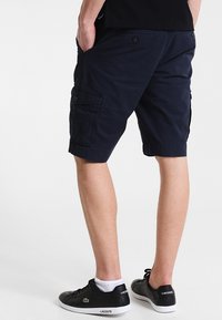 Uomo che indossa pantaloni cargo blu navy, sneakers Lacoste nere con suola bianca, calzini bianchi alla caviglia, in piedi con le mani nelle tasche.