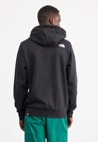 Zwarte hoodie met een wit logo op de achterkant, textuurstof en een ontspannen pasvorm, gecombineerd met groene cargobroeken.