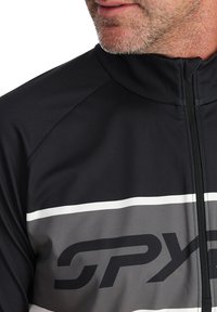 Giacca da ciclismo nera e grigia con chiusura a zip, caratterizzata da un logo prominente e due strisce bianche orizzontali sul petto.