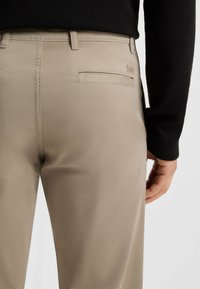 Pantaloni beige con una texture liscia, dotati di una tasca posteriore con etichetta del logo. Cuciture visibili e un design elegante e aderente.