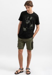 Zwarte katoenen T-shirt met een grafische print van een filmrol en projector; gecombineerd met olijfgroene cargo shorts en sandalen.