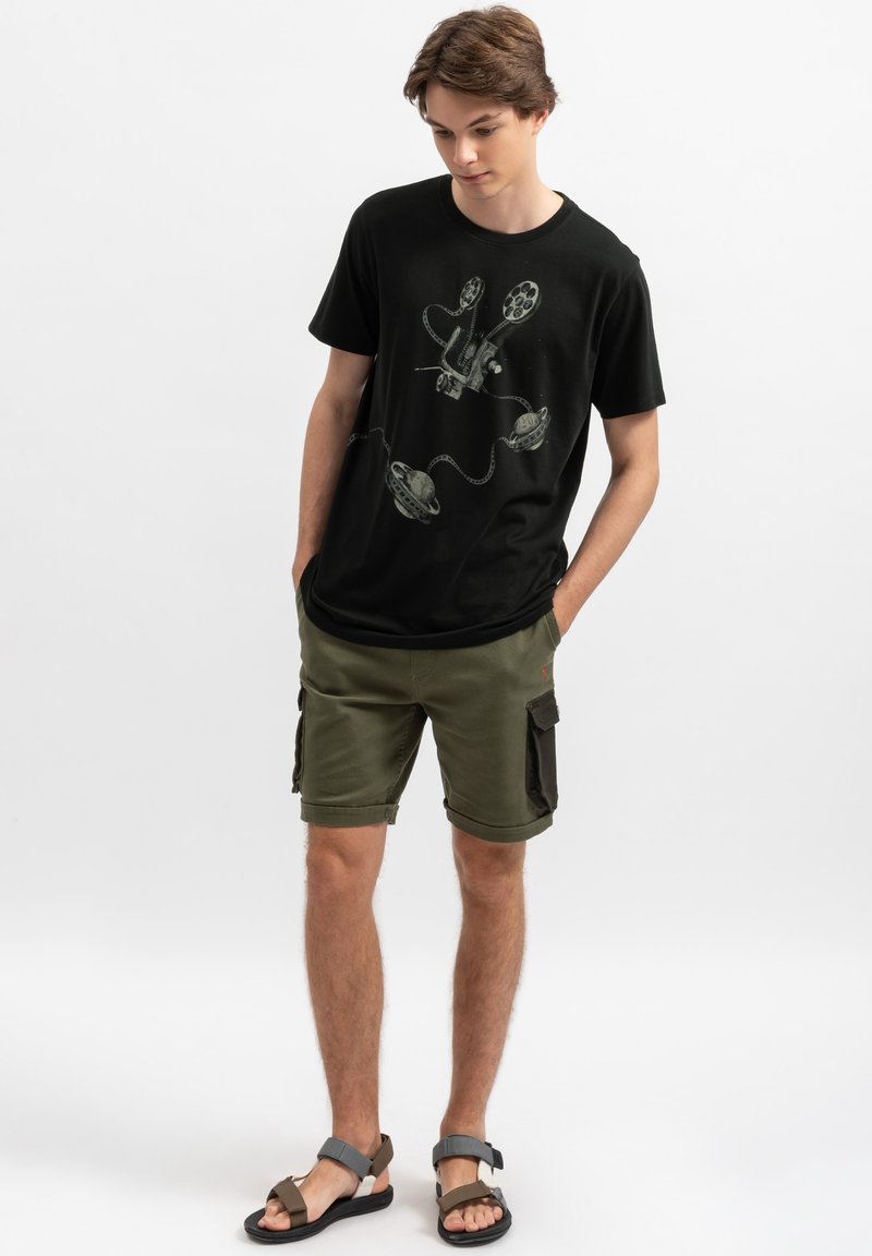 Zwarte katoenen T-shirt met een grafische print van een filmrol en projector; gecombineerd met olijfgroene cargo shorts en sandalen.