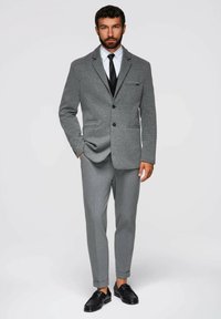 Grå strukturerad blazer med matchande grå skräddarsydda byxor. Svarta dressade skor accentuerar looken. Skjortan har en klassisk krage och slips.