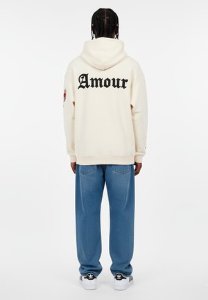 Felpa crema con "Amour" in lettere nere sul retro, abbinata a jeans larghi blu chiaro e sneaker bianche e nere.