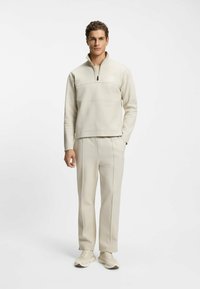 Μπεζ φλις μπλούζα με τέταρτο φερμουάρ, ραφή στο στήθος και λογότυπο· αντίστοιχο μπεζ παντελόνι με λεπτομέρειες ραφής στα πλάγια· συνδυασμένο με ανοιχτόχρωμα αθλητικά παπούτσια.