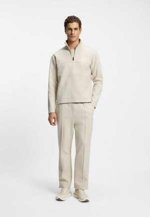 Beiger Fleecepullover mit Viertel-Reißverschluss, Brustnaht und Logo; passende beigen Hosen mit seitlicher Nahtführung; kombiniert mit hellen Sneakern.