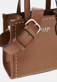 Borsa in pelle marrone con hardware in argento, dettagli cuciti e tracolla regolabile. Presenta il logo del marchio "GUESS EST1981" in lettere metalliche.
