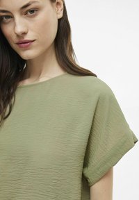 Šviesiai alyvų žalios spalvos trumpas rankovės viršus su apvaliu iškirpimu ir tekstūruotu audiniu, su suvyniotomis rankovėmis ir laisvos formos dizainu.