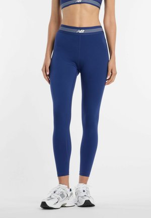 Frau trägt blaue Leggings mit hoher Taille und einen passenden Sport-BH mit weißen und blauen New Balance Sneakers auf weißem Hintergrund.