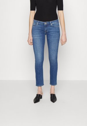 Kobieta ubrana w niebieskie obcisłe dżinsy, czarną krótką bluzkę z krótkim rękawem oraz czarne czółenka na obcasie z ostrym noskiem, stojąca na tle jednolitego jasnego tła.