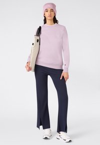 Felpa a girocollo viola chiaro, pantaloni svasati blu scuro con fessure laterali, abbinati a una borsa beige e sneakers bianche. Berretto di maglia in testa.