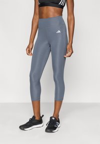 Γκρι, εφαρμοστά καπρί κολάν με ψηλή μέση, κατασκευασμένα από ελαστικό υλικό. Διαθέτουν λευκό λογότυπο Adidas και ορατές ραφές.