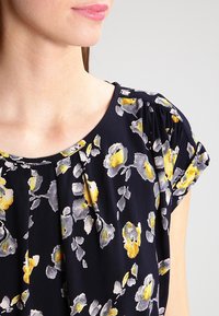 Haut noir à manches courtes avec un motif floral jaune et gris, comportant des plis au niveau du col, fabriqué en tissu léger.