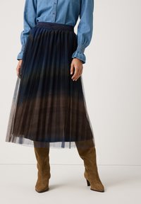 Der Tüllrock zeigt einen dunkelblauen bis braunen Farbverlauf, hat einen elastischen Bund und ein plissiertes Design. Kombiniert mit kniehohen braunen Wildlederstiefeln.