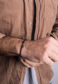 Black leather bracelet with decorative metal accents. Worn on a textured, hair-bearing wrist, paired with a brown corduroy shirt.