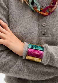 Maglione grigio in lana con tre braccialetti rettangolari: turchese, magenta e giallo. Tessuto morbido con dettagli a bottone sul colletto.