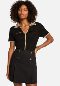 Polo en tricot noir avec col et accents beiges, présentant des manches courtes, une fermeture à boutons sur le devant et deux poches poitrine ; associé à une jupe noire.