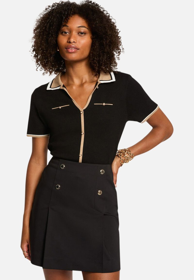 Polo en tricot noir avec col et accents beiges, présentant des manches courtes, une fermeture à boutons sur le devant et deux poches poitrine ; associé à une jupe noire.