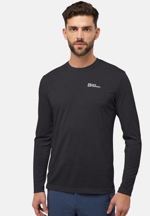 Man met kort bruin haar en baard, gekleed in een zwart Jack Wolfskin shirt met lange mouwen en een blauwe broek, staand tegen een effen achtergrond.
