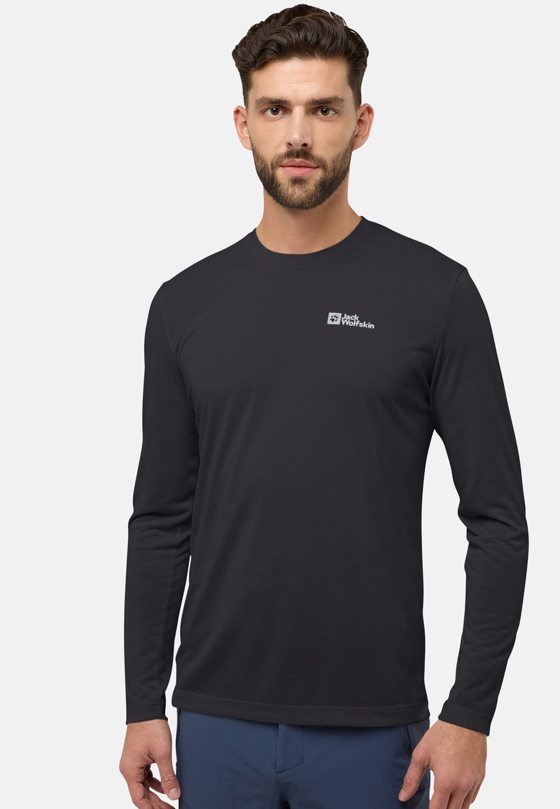 Mann mit kurzem braunem Haar und Bart, der ein schwarzes langärmliges Jack Wolfskin-Shirt und eine blaue Hose trägt, steht vor einem schlichten Hintergrund.