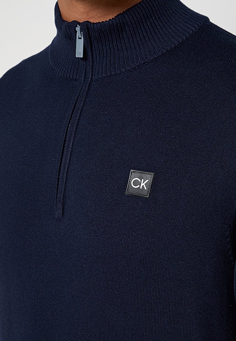 Pull en maille bleu marine avec un col haut et une fermeture éclair de quart. Présente une étiquette carrée noire avec "CK" sur le devant. Texture lisse.