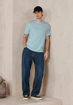 Jaunas vyras, dėvintis šviesiai mėlyną marškinėlį, tamsiai mėlynus džinsus, juodą kepurę ir juodus sportinius batelius, stovintis priešais smėlio spalvos sieną su viena ranka kišenėje.