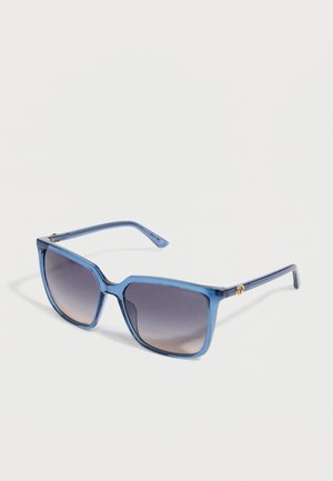 Blue translucent square-framed sunglasses with gradient dark lenses and golden logo detail on the temples resting on a white surface.