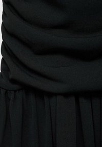 Crna haljina sa slojevitom tkaninom; sadrži naborani gornji deo i suknju s nabranim teksturom. Mekani pleteni materijal, profinjenog izgleda. Elegantan siluet.