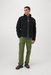Schwarze Pufferjacke mit hohem Kragen, kombiniert mit grünen Cargo-Hosen mit Seitentaschen. Modell trägt ein graues T-Shirt und mehrfarbige Sneakers.