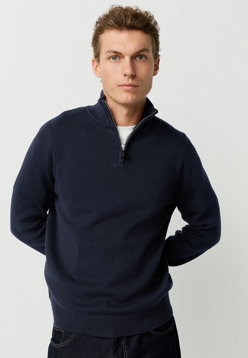 Jeune homme aux cheveux châtain clair portant un pull à demi-fermeture éclair bleu marine et un pantalon foncé, debout devant un fond clair uni.