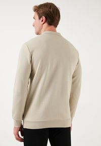 Pullover beige a maniche lunghe in tessuto strutturato, con colletto girocollo e polsini a costine, visto da dietro su uno sfondo semplice.