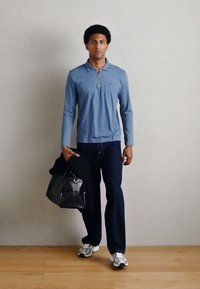 Hele sinine pikakäistega polomees, millel on tõmblukk, tumedad teksapüksid ja must käekott, kantud koos valgete ja hallide tossudega puidupõrandal.