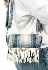 Geweven stoffen handtas met blauwe en beige strepen, voorzien van witte franjes aan de onderkant en een zilveren logo accent. Inclusief verstelbare strap.