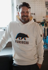 Sudadera blanca con capucha que presenta un gráfico de un oso en negro con acentos en azul y naranja, y la palabra "FORSBERG" abajo. Manga acampanada y bolsillo tipo canguro.