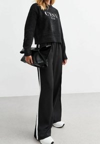 Sweat à capuche noir en peluche avec le texte "C'EST BON", associé à un pantalon noir ample avec des bandes latérales blanches. Sac à main noir texturé et baskets blanches.