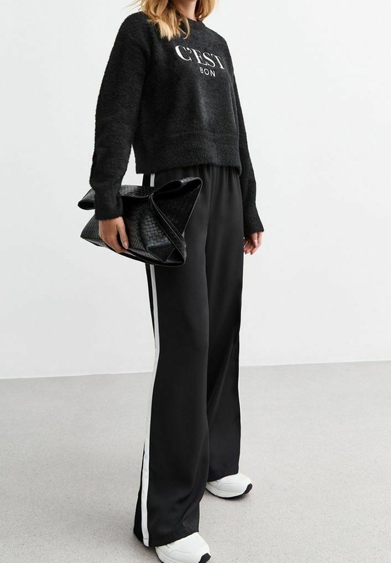 Sweat à capuche noir en peluche avec le texte "C'EST BON", associé à un pantalon noir ample avec des bandes latérales blanches. Sac à main noir texturé et baskets blanches.