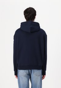 Sudadera con capucha de color azul marino en tela suave. Presenta un corte holgado, puños y dobladillo de canalé. La vista posterior muestra un diseño minimalista y la estructura de la capucha.
