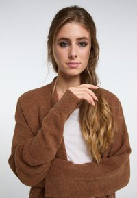 Rudas kardiganas, pagamintas iš minkštos medžiagos, su laisvu siluetu, ilgomis rankovėmis ir smulkiais megztiniais, dėvimas virš baltos marškinėlių.