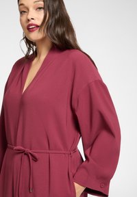 Bordowy szlafrok z dekoltem w serek, szerokimi rękawami i paskiem w talii. Wykonany z miękkiego materiału o gładkiej fakturze.