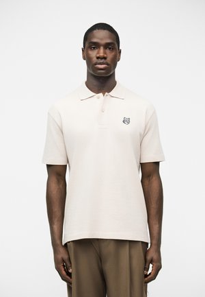 Noor mees kerge beežika lühikeste varrukatega polo särgiga, millel vasakul rinnal huntide logoga ja pruunide pükstega, seisab valge tausta ees.